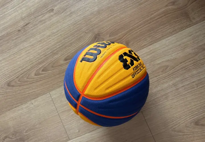 Piłka do koszykówki 3x3 Wilson z pomarańczowo-niebieskimi panelami leży na drewnianej podłodze.