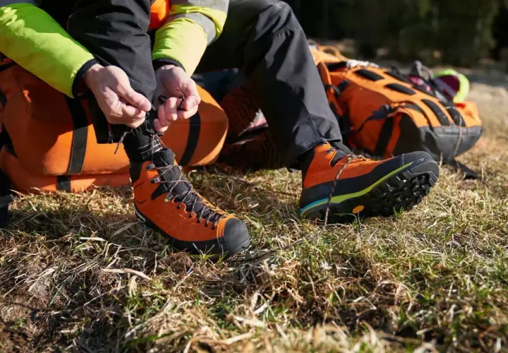 A person is lacing up bright orange hiking boots with a heel lock lacing system, preparing for an adventure with an orange backpack nearby.