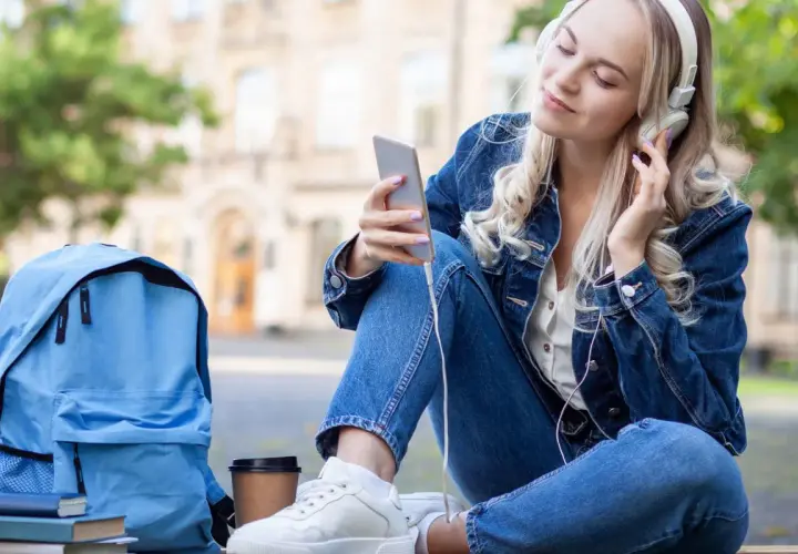 Dziewczyna w dżinsowej kurtce słucha muzyki na telefonie. Obok niej leży niebieski plecak i książki. Idealny moment, by pomyśleć, o czym można pisać z chłopakiem.