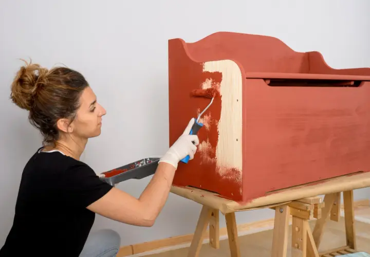 Mujer con guantes blancos pinta con rodillo una caja de madera de color terracota.
