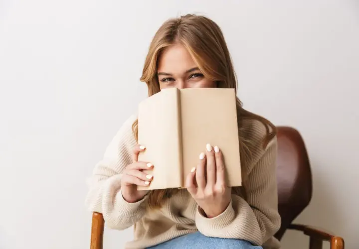 Dziewczyna z uśmiechem ukrywa twarz za książką, zanurzona w świecie literatury young adult.