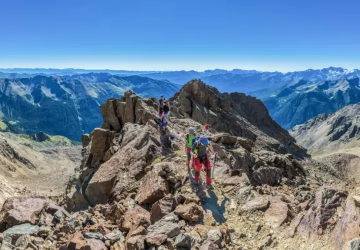 Grupa ludzi wyrusza na trekking po skalistym szczycie, podziwiając rozległe pasmo górskie w oddali.