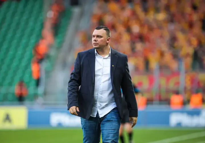 Trener Jagiellonii w eleganckim garniturze i dżinsach, na tle trybun stadionu wypełnionych kibicami.