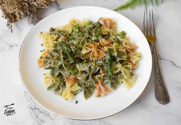 Plato de pasta primavera con farfalle tricolor, espárragos y guisantes en salsa cremosa, adornado con cebollino picado.