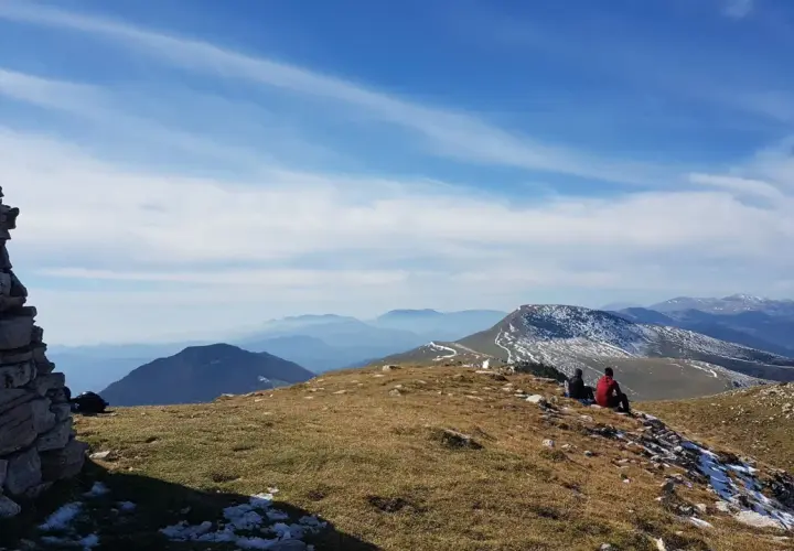 Dos personas contemplan el paisaje montañoso desde el Puig d'Estela, con un muro de piedra a la izquierda y el cielo azul con nubes.