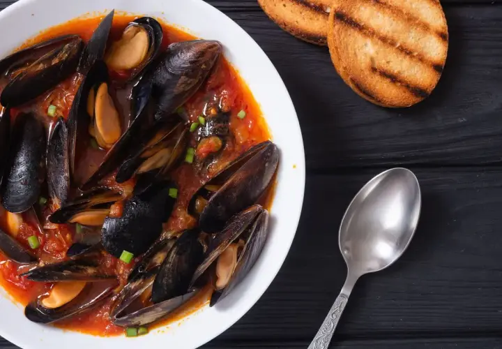 Un invitante piatto di zuppa di cozze in un ricco sugo di pomodoro, guarnito con erba cipollina, accompagnato da crostini tostati e un cucchiaio.