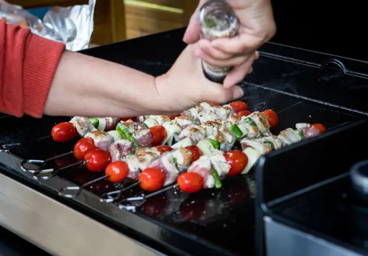 Brochettes de viande, tomates et courgettes sur une plancha à gaz. Une main assaisonne les brochettes.