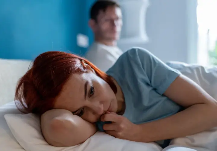 Smutna kobieta leży w łóżku, czując się samotna i przygnębiona. Jej partner jest w tle, ale nie okazuje wsparcia, co potęguje poczucie poniżania w związku.