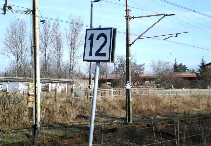 Wskaźnik W24 z numerem 12 przy torach kolejowych. W tle budynki i drzewa.
