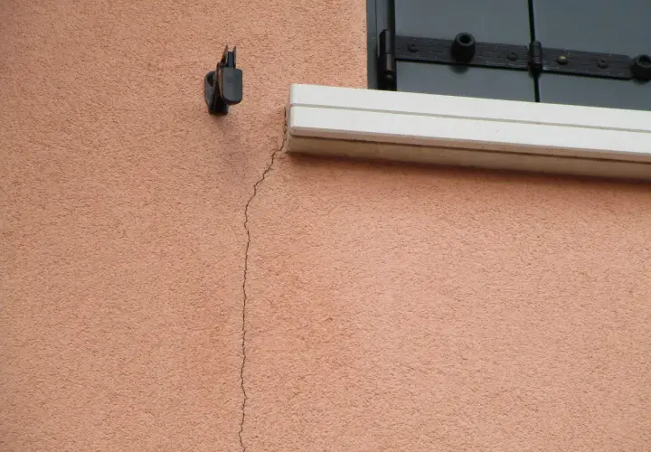 Una crepa sottile attraversa l'intonaco rosa di un muro esterno, vicino a un supporto metallico e a una finestra con persiana. Utile per capire come riempire le crepe nei muri esterni.
