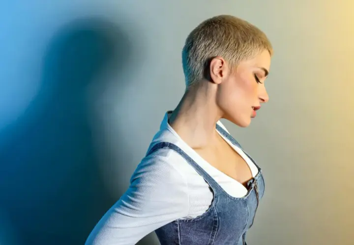 Kobieta z fryzurą buzz cut blond, w białej bluzce i dżinsowej sukience na szelkach, z profilu, z zamkniętymi oczami.