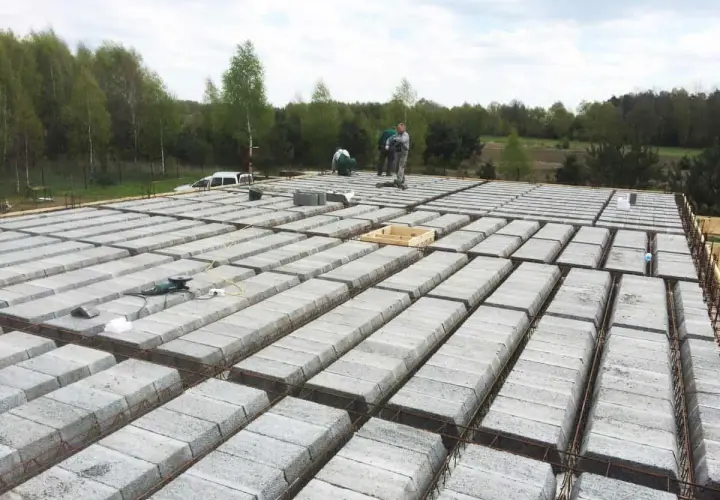 Budowa stropu teriva w toku. Betonowe pustaki ułożone w rzędach, zbrojenie gotowe do zalania. Pracownicy doglądają postępu prac.