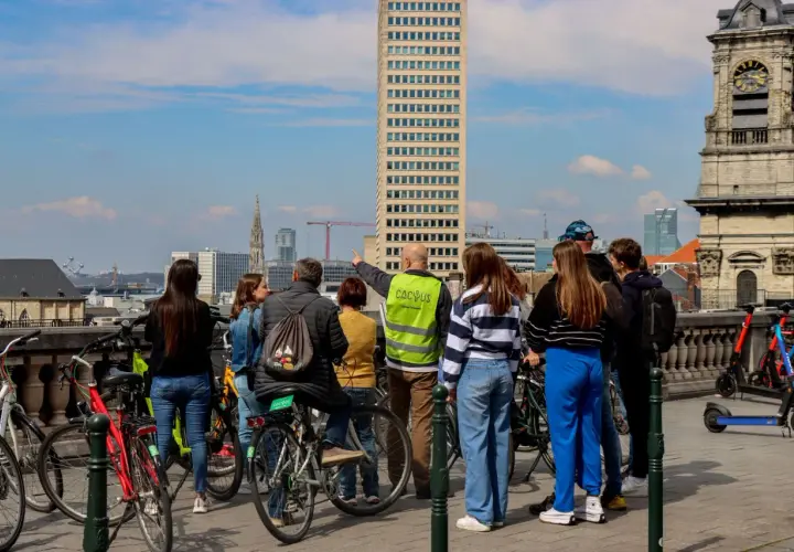 A group gathers for Brussels bike tours, with a guide pointing towards the city skyline featuring a tall skyscraper and an old clock tower.