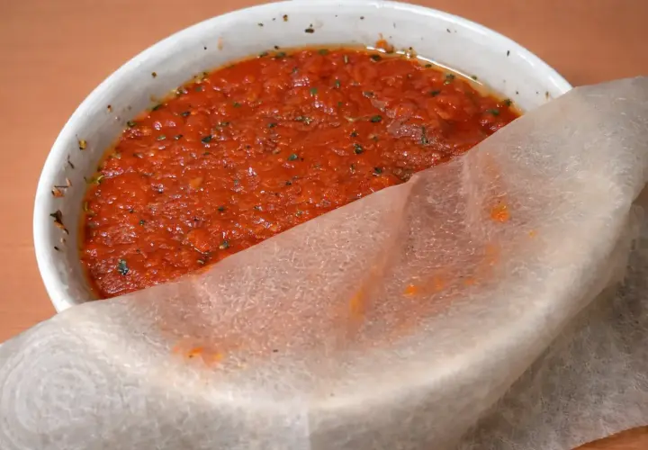 Un tazón blanco con la mejor salsa de tomate para pizza, con hierbas y un trozo de papel de arroz.