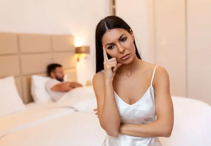 Donna pensierosa in camera da letto, mentre l'uomo è a letto. Potrebbe essere un momento per capire come aumentare la voglia di fare l'amore nell'uomo.