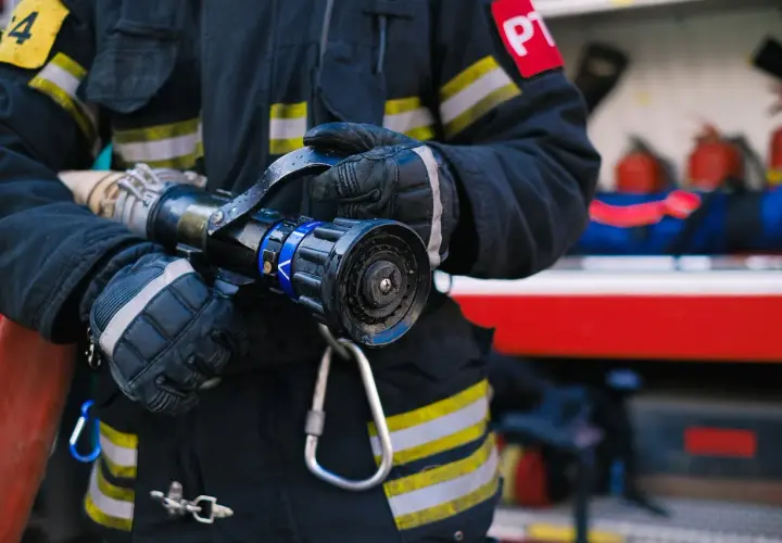 Strażak w rękawicach trzyma prądownicę. Zastanawiasz się, jakie są stopnie w PSP po ilu latach?
