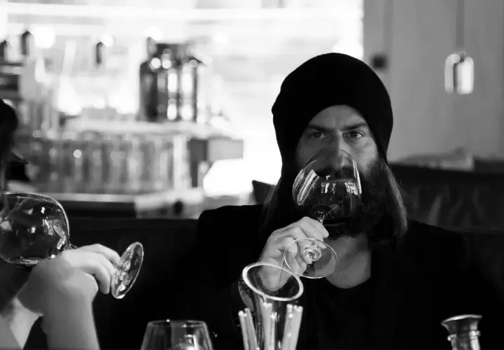 Ein Mann mit Bart und Mütze trinkt Wein. Er hält das Glas am Stiel, eine elegante Art, wie trinkt man wein.