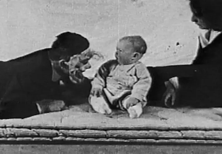 Bambino seduto, un uomo con maschera e una donna sullo sfondo, scena dall'esperimento piccolo Albert.