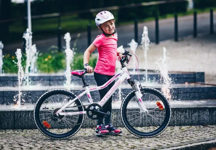 Uśmiechnięta dziewczynka w kasku stoi obok swojego białego roweru dla 7 latki, z fontannami w tle.