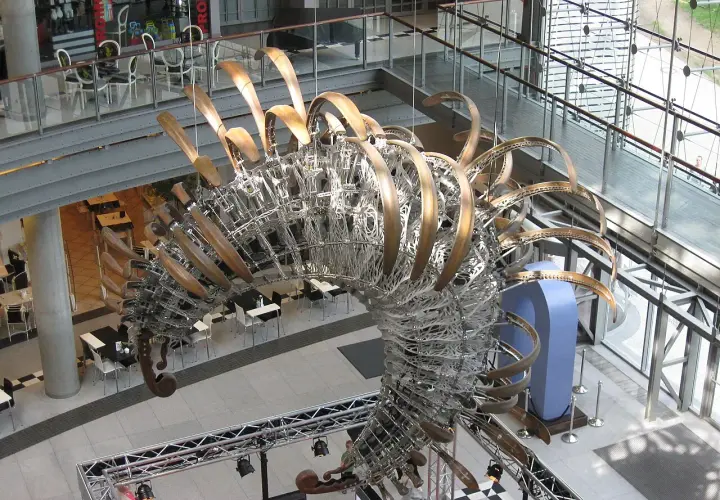 Duża, metalowa rzeźba przypominająca szkielet dinozaura w centrum handlowym.