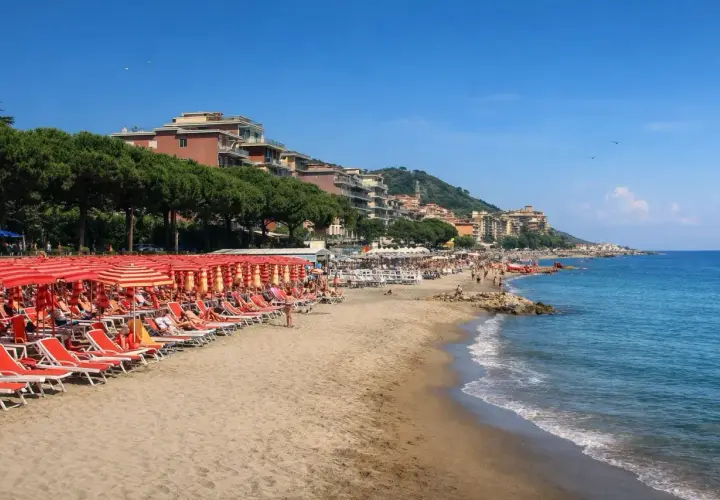 Plage d'Imperia animée, avec des rangées de parasols rouges et des chaises longues sur le sable. La mer bleue étincelle sous un ciel clair.