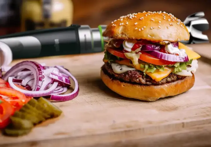 Pyszny burger z serem, warzywami i domowym sosem do burgera. Obok pokrojona cebula, pomidor i ogórek.