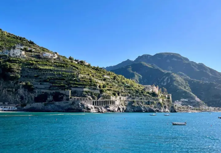 A scenic view from a Naples to Amalfi Coast ferry, showing terraced hillsides dotted with houses, a grand villa, and the sparkling blue sea.