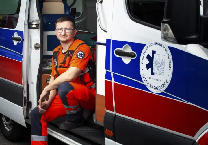 Mężczyzna w pomarańczowym stroju ratownika medycznego siedzi w otwartych drzwiach karetki.