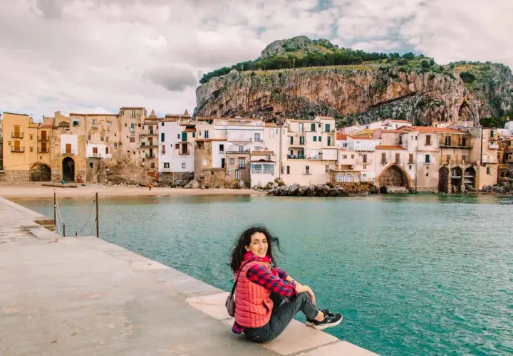 A woman smiles, enjoying her first time visiting Sicily. She sits on a stone pier overlooking the turquoise sea and a picturesque town with a dramatic cliff behind it.