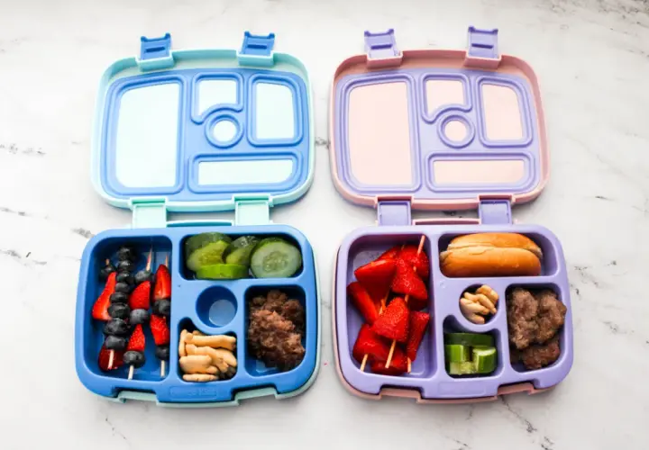 Two bento boxes, one blue and one purple, are filled with lunch items. The blue box contains fruit skewers, cucumber slices, crackers, and ground meat. The purple box holds strawberry skewers, a bun, nuts, and cucumber slices.