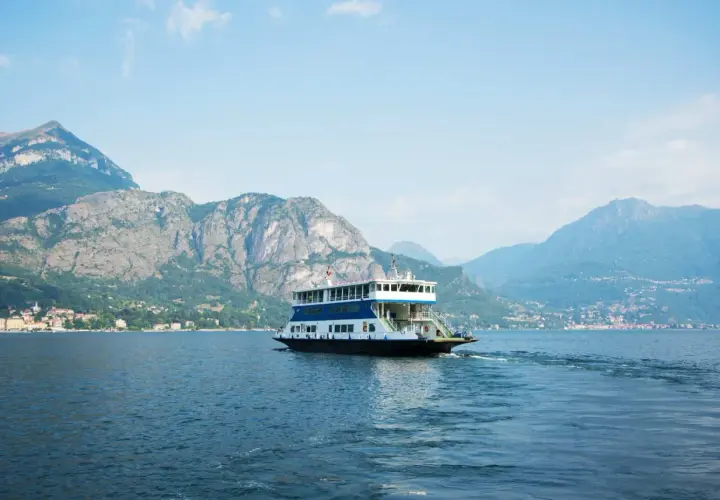Un ferry transporte des voitures sur le lac de Côme, naviguant paisiblement entre des montagnes majestueuses sous un ciel bleu.