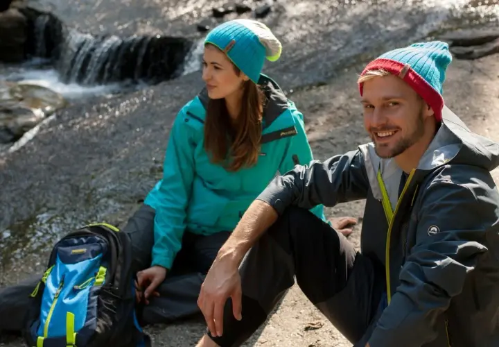 Para, co to jest trekking? Para w czapkach siedzi przy strumieniu, obok plecak.