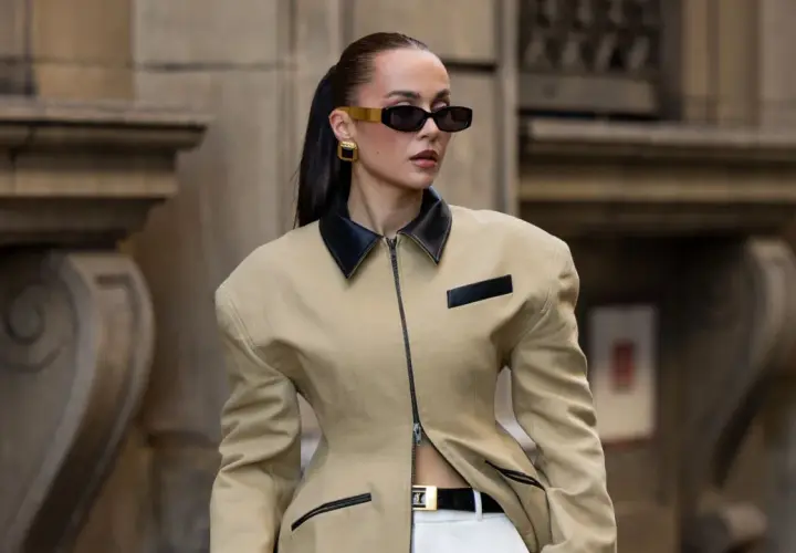 Mujer con gafas de sol oscuras y pendientes dorados, vistiendo una chaqueta beige con cuello negro y cinturón. Un look muy chic.