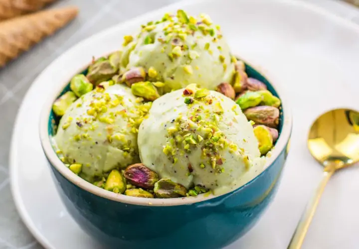 Tre palline di cremoso gelato al pistacchio, guarnite con granella e pistacchi interi, in una ciotola blu.