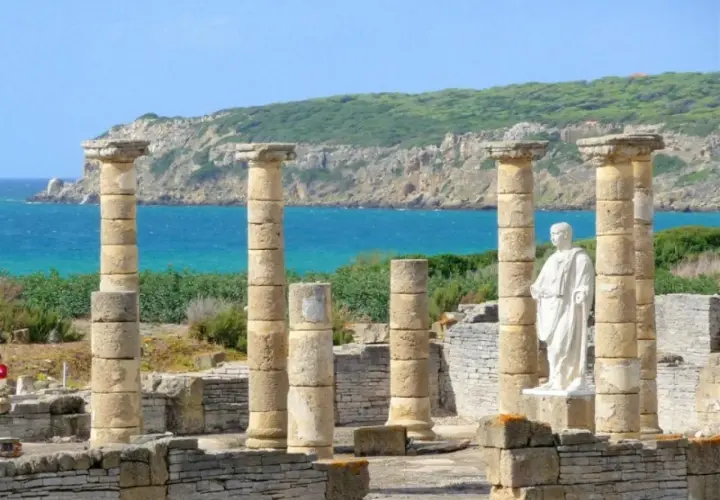 Ruinas romanas en el área de Bolonia, con columnas de piedra y una estatua blanca frente al mar azul y una colina verde.