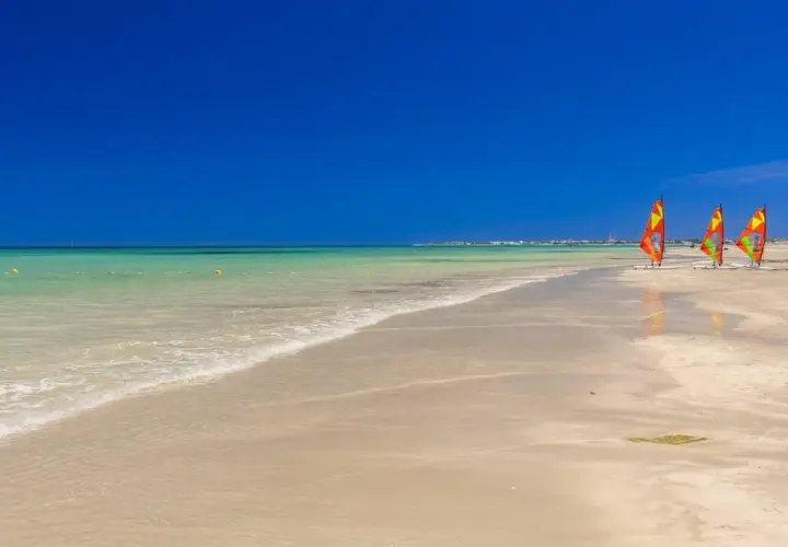 Piękna plaża w Tunezji w czerwcu. Trzy kolorowe deski windsurfingowe stoją na piasku, a turkusowa woda łagodnie faluje.