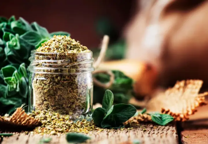 Ein Glas mit getrocknetem Oregano steht auf einem Holztisch. Frische Oreganozweige und weitere Gewürze sind im Hintergrund zu sehen.
