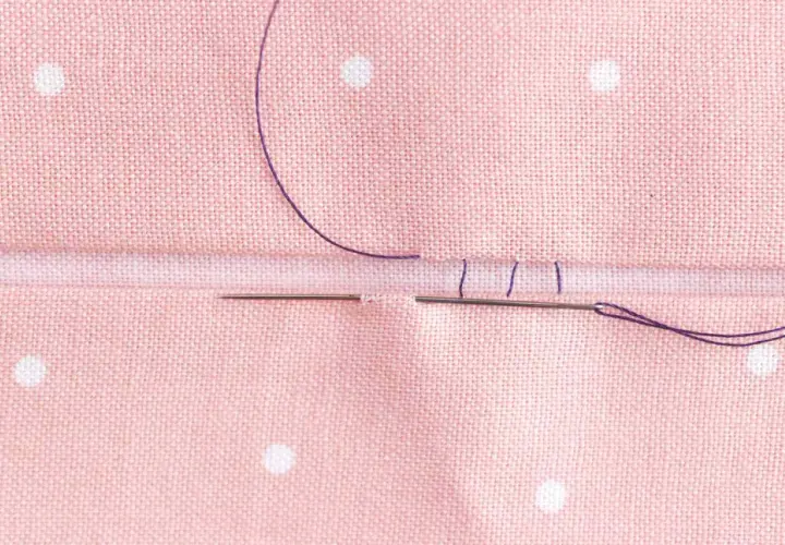 Close-up of a needle and purple thread creating an invisible hem stitch on pink polka-dot fabric.