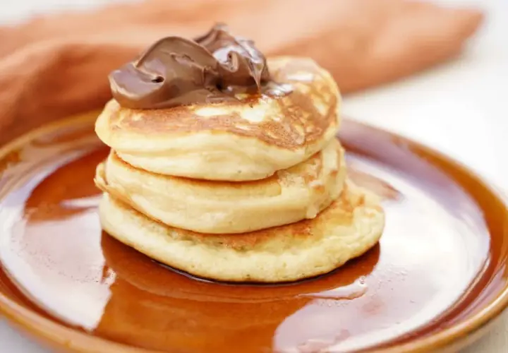 En stapel fluffiga amerikanska pannkakor toppas med chokladkräm på en brun tallrik.