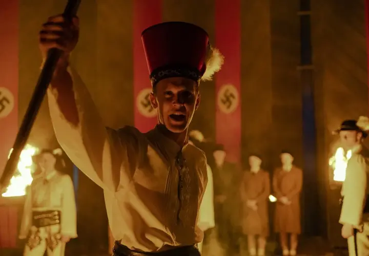 Młody mężczyzna w czerwonym nakryciu głowy z piórem wznosi płonący kij. W tle widać flagi ze swastykami, sugerujące scenę z filmu "Biała odwaga".