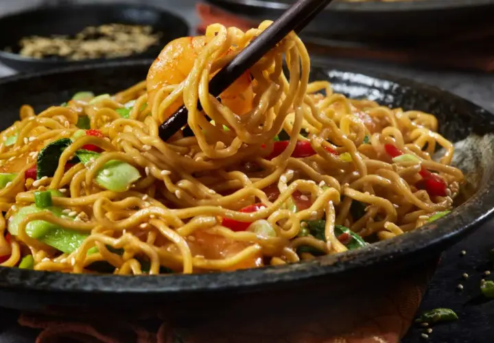 Un plato humeante de fideos con pollo, salteado con verduras y semillas de sésamo, listo para ser disfrutado.