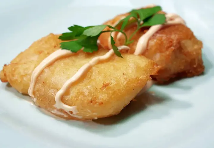 Dos trozos de bacalao rebozado, dorados y crujientes, servidos con una salsa cremosa y perejil fresco.