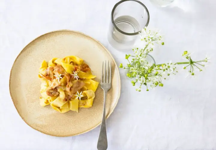Ein Teller mit cremiger Pasta, die wie Fettuccine aussieht, garniert mit zarten Blüten und Speck. Daneben ein Glas Wasser und ein Strauß weißer Blumen.
