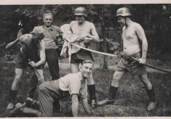 Fünf Männer, einige mit Helmen, posieren mit Waffen. Ein Mann hält eine Puppe. Die Szene erinnert an die Verbrechen der Wehrmacht.