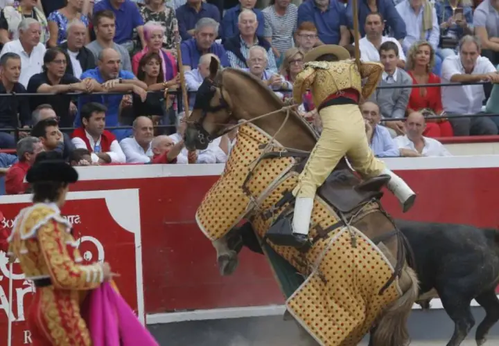 Un rejoneador, quizás Ana Romero, monta un caballo cubierto con un capote mientras un toro se acerca.