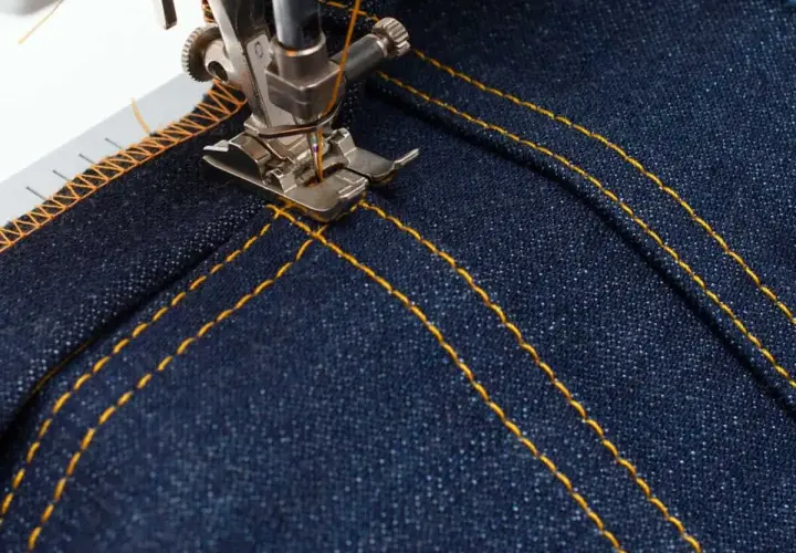 A sewing machine's topstitching foot creates neat, parallel yellow stitches on dark denim fabric, adding a professional finish.
