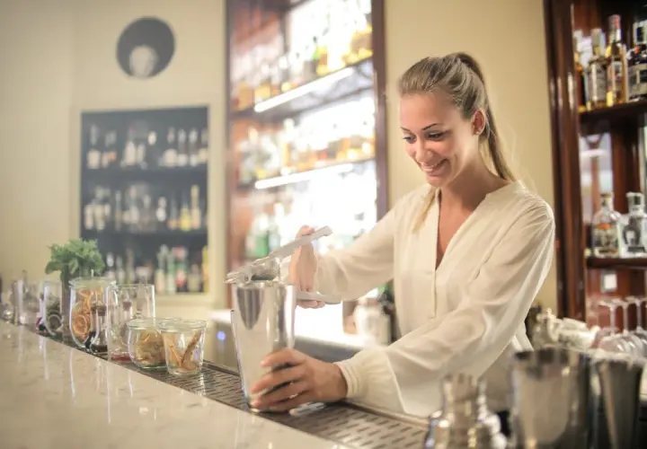 Uśmiechnięta barmanka przygotowuje drinka, ucząc się, jak zostać barmanem. Za nią półki z alkoholem.