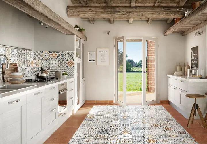 Cucina rustica con pavimento decorato, pensili bianchi e un'ampia porta finestra che si apre su un prato verde. La posa battiscopa è curata nei dettagli.