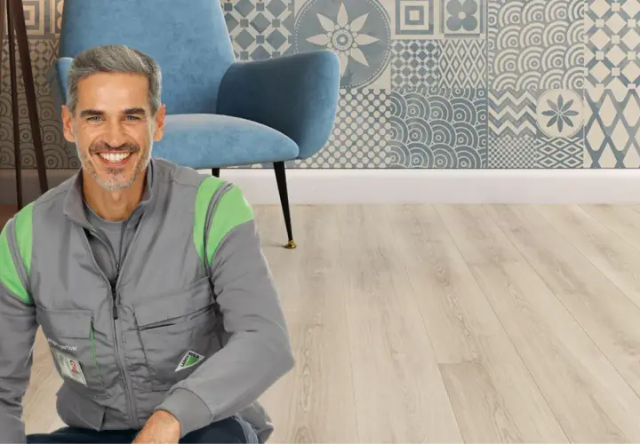 Un uomo sorridente in gilet da lavoro, pronto per il montaggio SPC. Sfondo con poltrona blu e parete decorata.