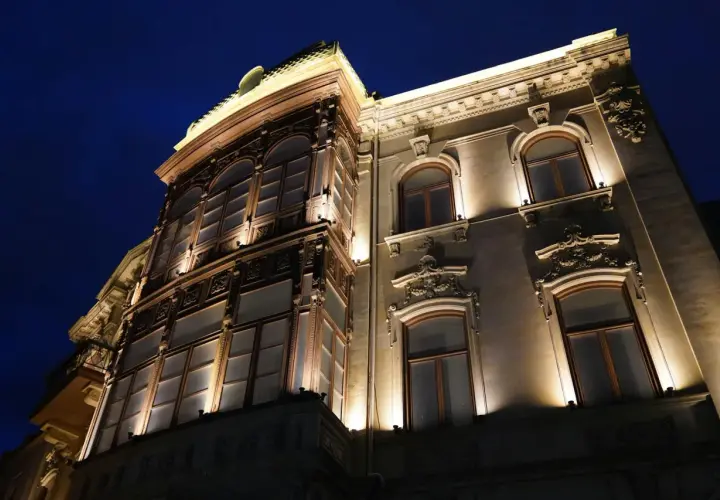 Elegancka kamienica nocą, oświetlona ciepłym blaskiem lamp miejskich. Widać bogate zdobienia fasady i duże, łukowe okna.